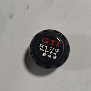 May include: Black shift knob with the word "GTI" in red and white lettering. The shift pattern is also in white. The knob has a round shape with small indentations around the edge.
