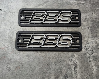 Può includere: Due emblemi rettangolari in plastica nera con un logo "BBS" bianco. Gli emblemi hanno un motivo a griglia.