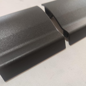 Puede incluir: Dos piezas de moldura de plástico negro para un automóvil. Las piezas son texturizadas y tienen una forma ligeramente curvada.