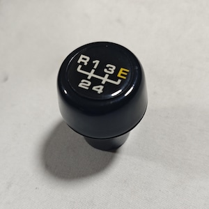 Puede incluir: Pomo de cambio de marchas negro con letras blancas que muestran el patrón de marchas: R, 1, 3, E, 2, 4.