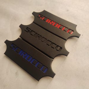 Puede incluir: Tres placas metálicas negras con la palabra "SCIROCCO" en rojo, blanco y azul. Las placas tienen un acabado texturizado y una forma distintiva con bordes curvos. Accesorios para automóviles.