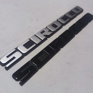 May include: Two car emblems, one silver and one black, both with the word "SCIROCCO" embossed on them.