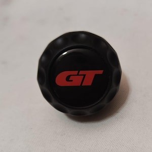 Peut inclure: Pièce automobile noire et circulaire avec le logo "GT" rouge. La pièce a un bord extérieur texturé et strié et un centre brillant et réfléchissant. Le logo est en gras, police sans-serif.