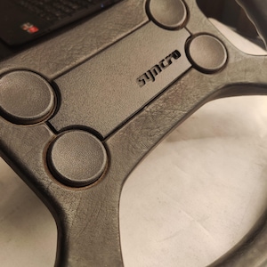 Può includere: Primo piano di un volante nero con la scritta "syncro" in rilievo al centro. Il volante ha una superficie strutturata e quattro pulsanti circolari. Il design è semplice e funzionale, probabilmente per un veicolo.