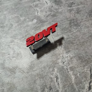 Könnte beinhalten: Rotes Plastiketikett mit dem Text "20VT" in einem fetten, 3D-Stil. Das Etikett ist auf einer kleinen schwarzen Basis montiert.