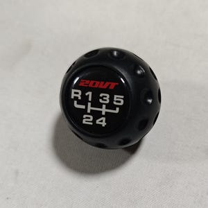 Peut inclure: Pommeau de levier de vitesses noir avec un motif circulaire et un schéma de changement de vitesse. Le bouton porte le texte "20VT" en rouge et le schéma de vitesses "R 1 3 5" et "2 4" en blanc. Le bouton a de petites fossettes.