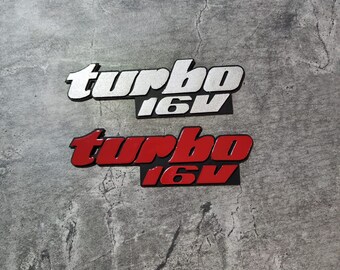 Może przedstawiać: Dwa metalowe emblematy samochodowe, jeden srebrny, a drugi czerwony, oba z napisem "turbo 16v".