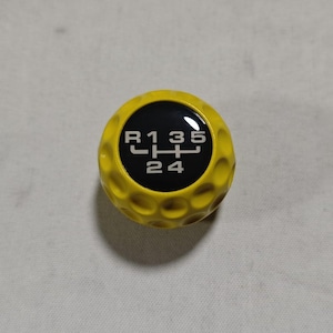 Könnte beinhalten: Ein leuchtend gelber Schaltknauf mit einer genoppten Textur, die an einen Golfball erinnert. In der Mitte befindet sich eine schwarze, kreisförmige Einlage mit einem weißen Schaltbild für ein Schaltgetriebe mit den Gängen R, 1, 2, 3, 4 und 5.