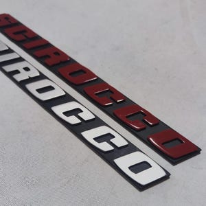Puede incluir: Dos emblemas de coche negros y rojos con la palabra "Scirocco" en letras blancas.