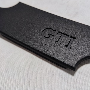 Könnte beinhalten: Schwarzes Kunststoff-Autoteil mit dem eingeprägten Wort "GTI".