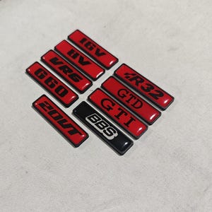 Op de afbeelding: Een verzameling rechthoekige auto-emblemen in rood en zwart. De emblemen tonen verschillende teksten, waaronder "16V", "VR6", "G60", "20VT", "R32", "GTD", "GTI" en "BBS". De emblemen zijn gerangschikt op een lichtgekleurd oppervlak.