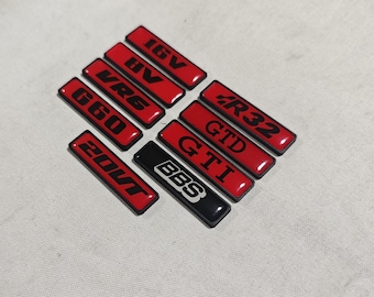 May include: A collection of rectangular car badges in red and black. The badges feature various text including "16V", "VR6", "G60", "20VT", "R32", "GTD", "GTI", and "BBS". The badges are arranged on a light-colored surface.