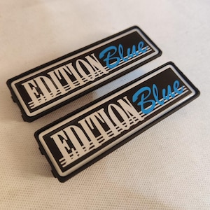 Könnte beinhalten: Zwei schwarze rechteckige Autoembleme mit silberner Einfassung und weißen Buchstaben. Der Text auf den Emblemen lautet "EDITION Blue" in blauen Buchstaben.