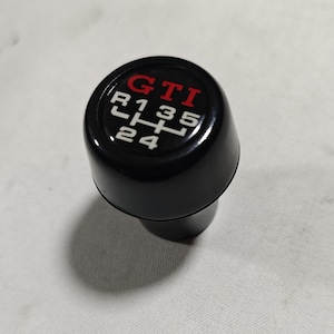 May include: Black gear shift knob with a red "GTI" logo and white numbers and letters indicating gear positions: R, 1, 3, 5, 2, 4.