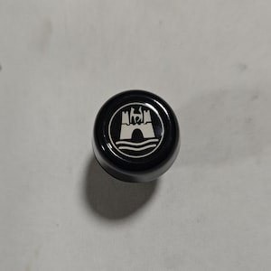 Op de afbeelding: Een zwarte plastic knop met een zilveren Volkswagen-logo met een kasteel en golven.