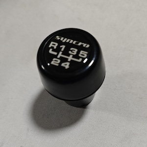 Könnte beinhalten: Schwarzer Schaltknauf mit weißer Beschriftung "Synchro R135 24".