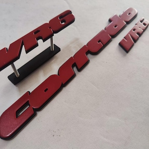 Może przedstawiać: Czerwone plastikowe emblematy samochodowe "WR6" i "Contrado". Emblemat "WR6" jest zamontowany na czarnej plastikowej podstawie.