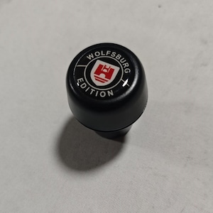 May include: A black gear shift knob featuring a circular emblem on top. The emblem displays the text WOLFSBURG EDITION in white lettering surrounding a red and white crest with a castle icon. The knob has a smooth, rounded, matte black finish.