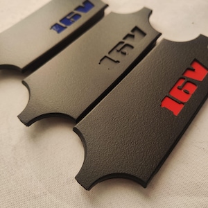Op de afbeelding: Vier donkergrijze rechthoekige auto-emblemen met een getextureerd oppervlak. Elk embleem heeft een unieke kleur voor de tekst "16V": blauw, zwart en rood. De emblemen hebben een kenmerkende vorm met gebogen randen.