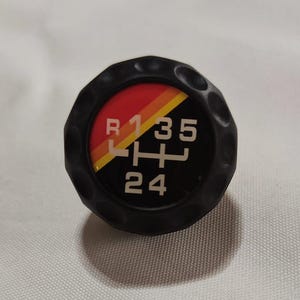 Peut inclure: Pommeau de levier de vitesses noir avec un design à rayures rouge, jaune et orange. Le pommeau comporte des chiffres blancs 1, 3, 5, 2 et 4, ainsi que la lettre R.