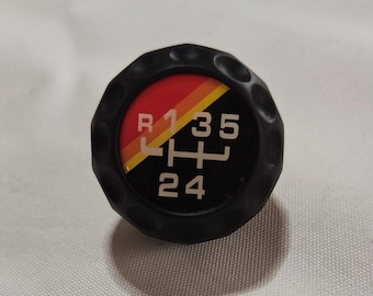 Può includere: Pomello del cambio nero con un design a strisce rosso, giallo e arancio. Il pomello ha i numeri bianchi 1, 3, 5, 2 e 4 e la lettera R.