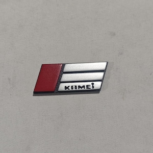 May include: A rectangular emblem with a red section on the left and a silver section with three horizontal lines. The word "KAMEI" is printed in black on the silver section. The emblem is on a gray surface.