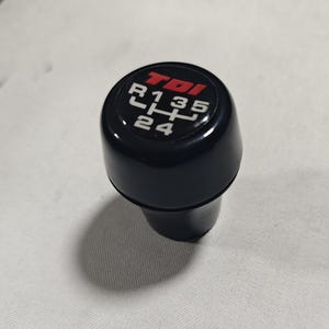 Peut inclure: Pommeau de levier de vitesses noir avec des chiffres blancs et un logo "TDI" rouge. Les chiffres sur le pommeau sont "R 1 3 5 4 2".