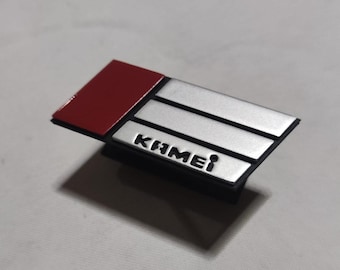 Può includere: Un cerotto rettangolare nero e argento con la parola "KAMEI" in lettere bianche. Il cerotto ha una striscia rossa nella parte superiore.