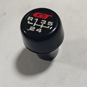 May include: Black gear shift knob with a red "GT" logo and white numbers 1, 2, 3, 4, 5, and R.