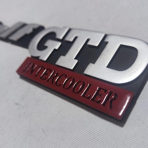 May include: Silver and black car emblem with the text "GT Intercooler" in red.