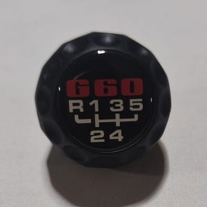 Pode incluir: Botão de câmbio preto com um padrão de 6 velocidades vermelho e branco. O botão tem uma superfície texturizada e os números "660" são impressos em vermelho.