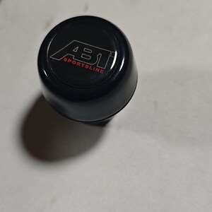 Puede incluir: Un objeto cilíndrico negro con el texto "ABT SPORTSLINE" en blanco y rojo. El objeto tiene un acabado brillante, posiblemente una pieza o accesorio de automóvil.