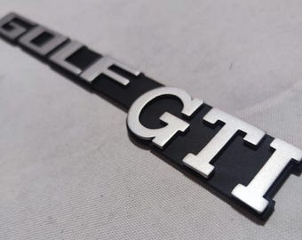 May include: A black and silver car emblem with the words "GOLF GTI" in raised silver lettering.