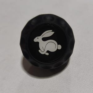 Peut inclure: Une balle de golf noire avec un logo de lapin blanc. Le lapin est tourné vers la droite et a de longues oreilles.