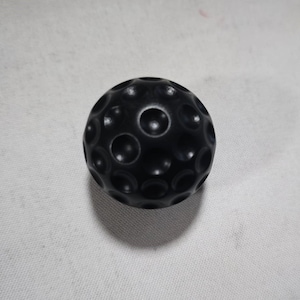 Könnte beinhalten: Ein schwarzer, kugelförmiger Gegenstand mit einer strukturierten Oberfläche, die zahlreiche kreisförmige Vertiefungen aufweist. Das Objekt hat eine einfarbige Farbe und scheint aus einem glatten Material zu bestehen. Das Design erinnert an einen Golfball.