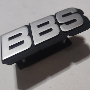 Könnte beinhalten: Ein silbernes BBS-Logo auf einem schwarzen Kunststoffsockel mit zwei Schrauben.