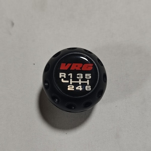 以下が含まれることがあります： 赤色の「VRG」の文字と白いギアシフトパターンが特徴の黒いギアシフトノブ。ノブは丸い形で、側面に小さな円形のくぼみがあります。