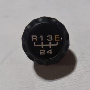 Puede incluir: Pomo de cambio de marchas negro con letras blancas que dicen "R13E" y "24" con un patrón de cambio de marchas blanco.