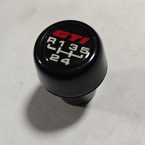 Peut inclure: Pommeau de levier de vitesses noir avec un logo "GTI" rouge et des chiffres blancs 1, 2, 3, 4, 5 et R.