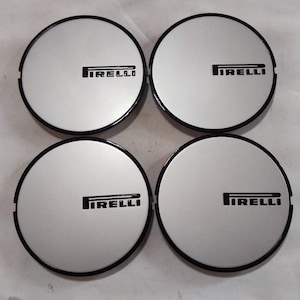 May include: A set of four circular wheel center caps featuring a silver metallic finish and a black border. Each cap displays the black Pirelli logo printed horizontally across the center. The design is consistent across all four individual pieces.