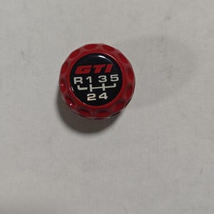 May include: A red GTI shift knob with a black center. The black center displays the GTI logo and the gear shift pattern in white. The knob has a textured surface.