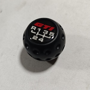 Op de afbeelding: Zwarte versnellingspookknop met de letters "GTI" in rood en wit, en een schakelpatroon. De knop heeft een geribbeld oppervlak.