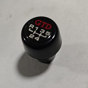 May include: Black gear shift knob with white numbers and a red "GTD" logo.