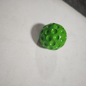 Puede incluir: Una pelota de golf verde brillante con una superficie con hoyuelos. La pelota es de un color verde sólido y vibrante, con numerosas pequeñas hendiduras circulares en su superficie. La pelota es de tamaño estándar para el deporte.