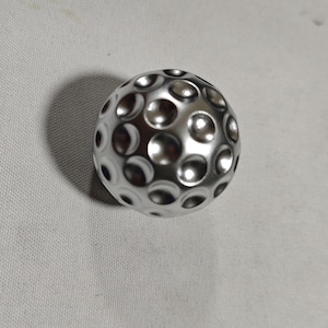 Op de afbeelding: Een zilverkleurig object in de vorm van een golfbal met talrijke putjes. De metalen bal reflecteert licht, waardoor highlights en schaduwen ontstaan. De putjes zijn gelijkmatig verdeeld.