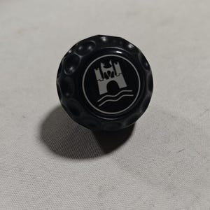 May include: A black spherical knob with a dimpled, golf ball-like texture. The center features a circular emblem displaying a white graphic of a castle tower positioned above wavy lines, all enclosed within a thin white circular border.
