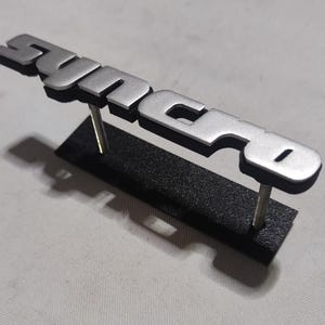 Può includere: Un emblema in metallo argentato con la parola "syncro" in lettere in rilievo. L'emblema è montato su un supporto nero.