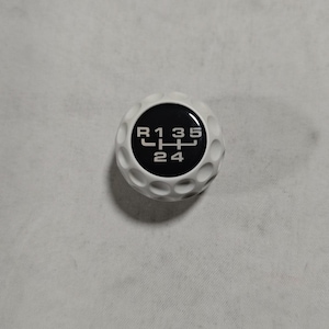 Può includere: Un pomello del cambio sferico bianco con una texture a fossette che ricorda una pallina da golf. Il centro presenta un inserto circolare nero che mostra uno schema di trasmissione manuale bianco con le posizioni R, 1, 2, 3, 4 e 5.
