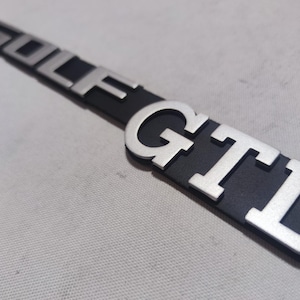 Op de afbeelding: Een zilveren "GOLF GTI" embleem met een zwarte achtergrond. De letters zijn verhoogd en hebben een gestructureerd oppervlak.