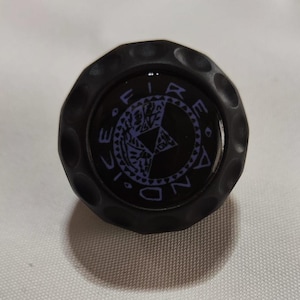 Op de afbeelding: Ronde, zwarte, getextureerde knop met een zwart en paars Fire and Ice-logo.